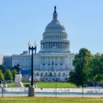 The United States Capitol Building, a white domed structure, stands prominently against a clear blue sky, surrounded by green lawns and trees, with a lamppost and statues in the foreground.