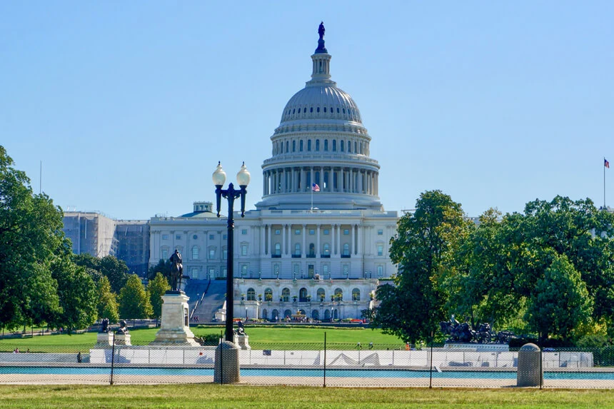 The United States Capitol Building, a white domed structure, stands prominently against a clear blue sky, surrounded by green lawns and trees, with a lamppost and statues in the foreground.